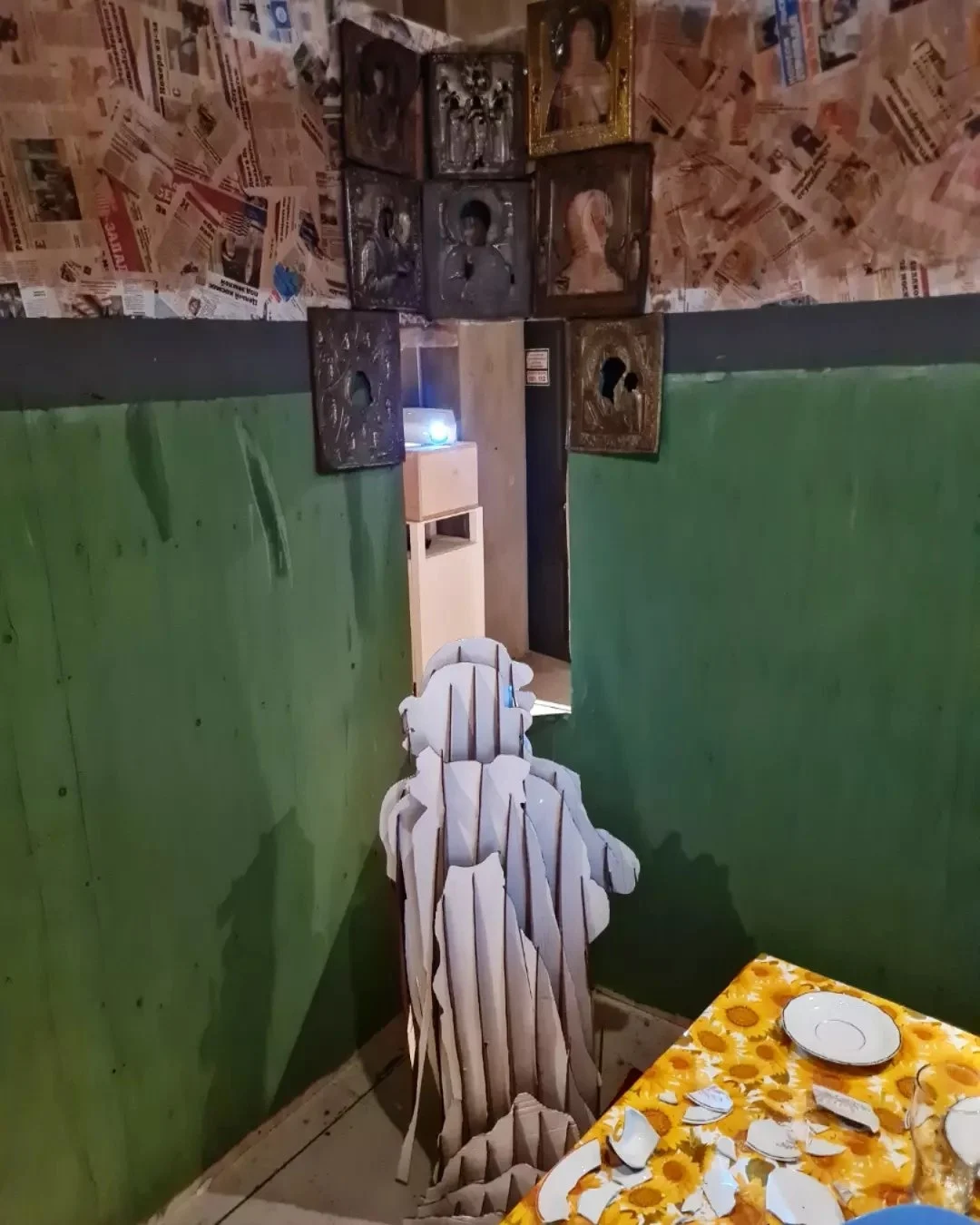 Icons on newspaper-covered walls, cardboard figure